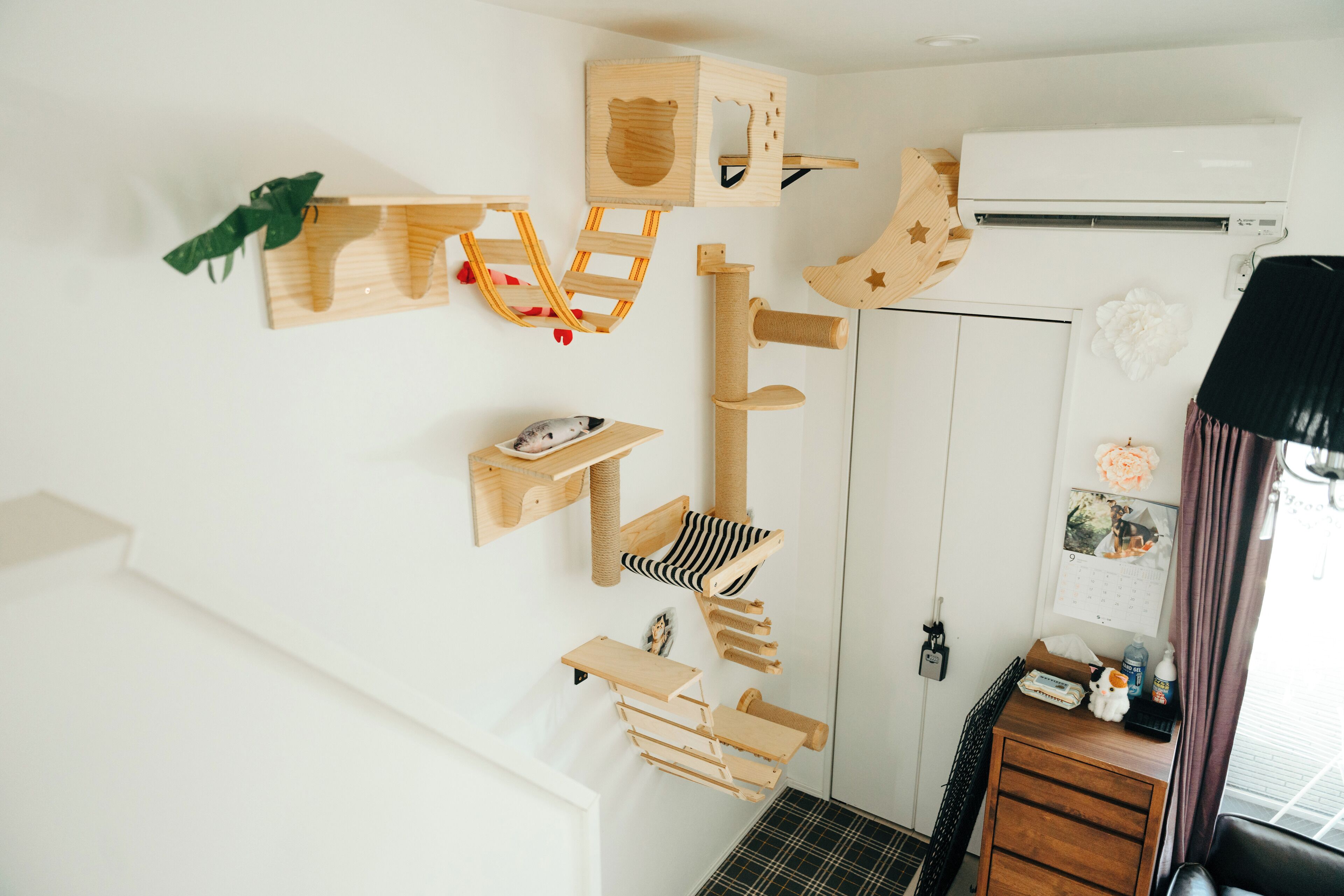 A room with a wooden cat tree, a calendar, a small desk with a chair, and a window with curtains.