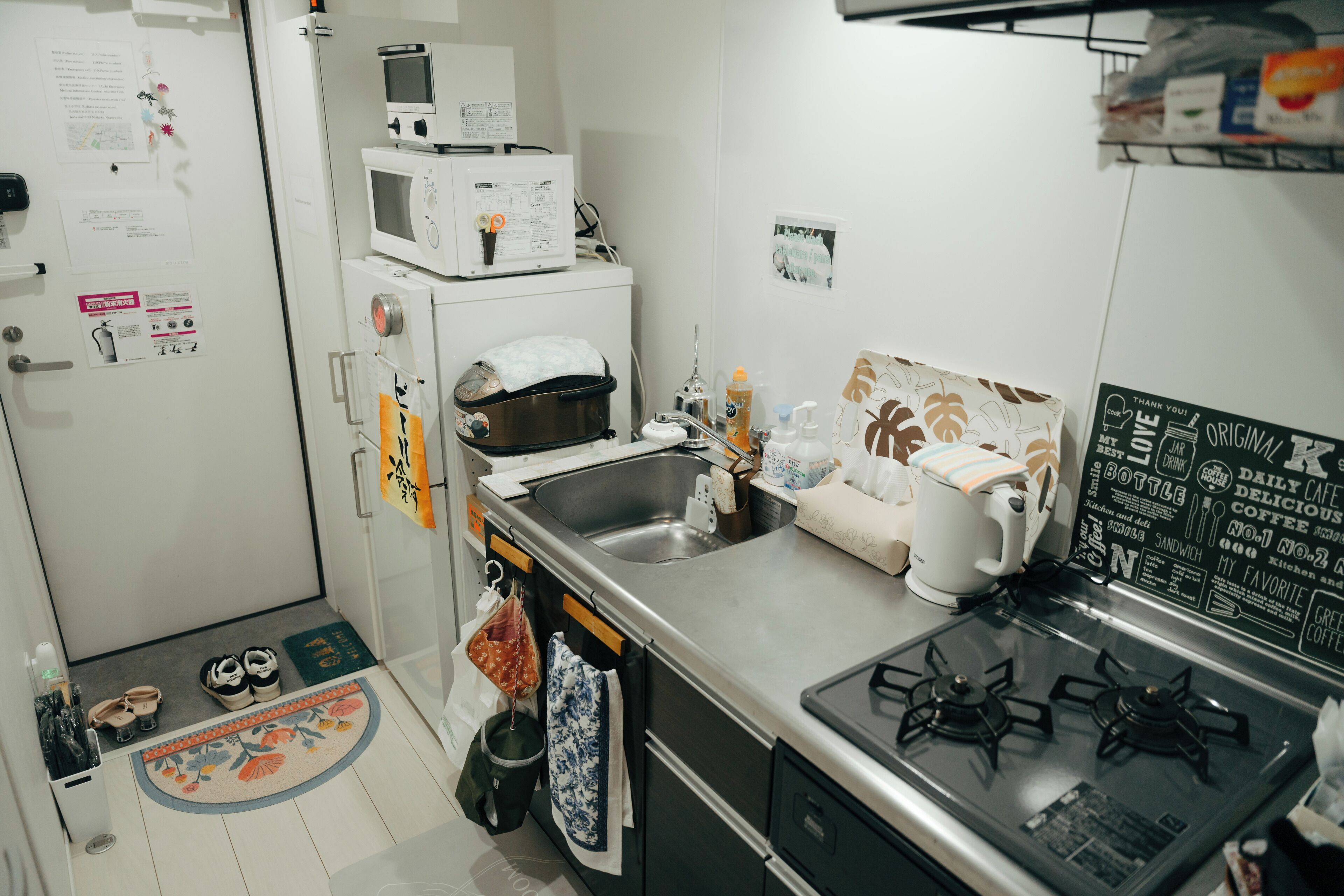Cocina compacta con microondas, estufa y lavabo. Zapatos de niño cerca de la puerta, un cesto de basura pequeño y un felpudo.