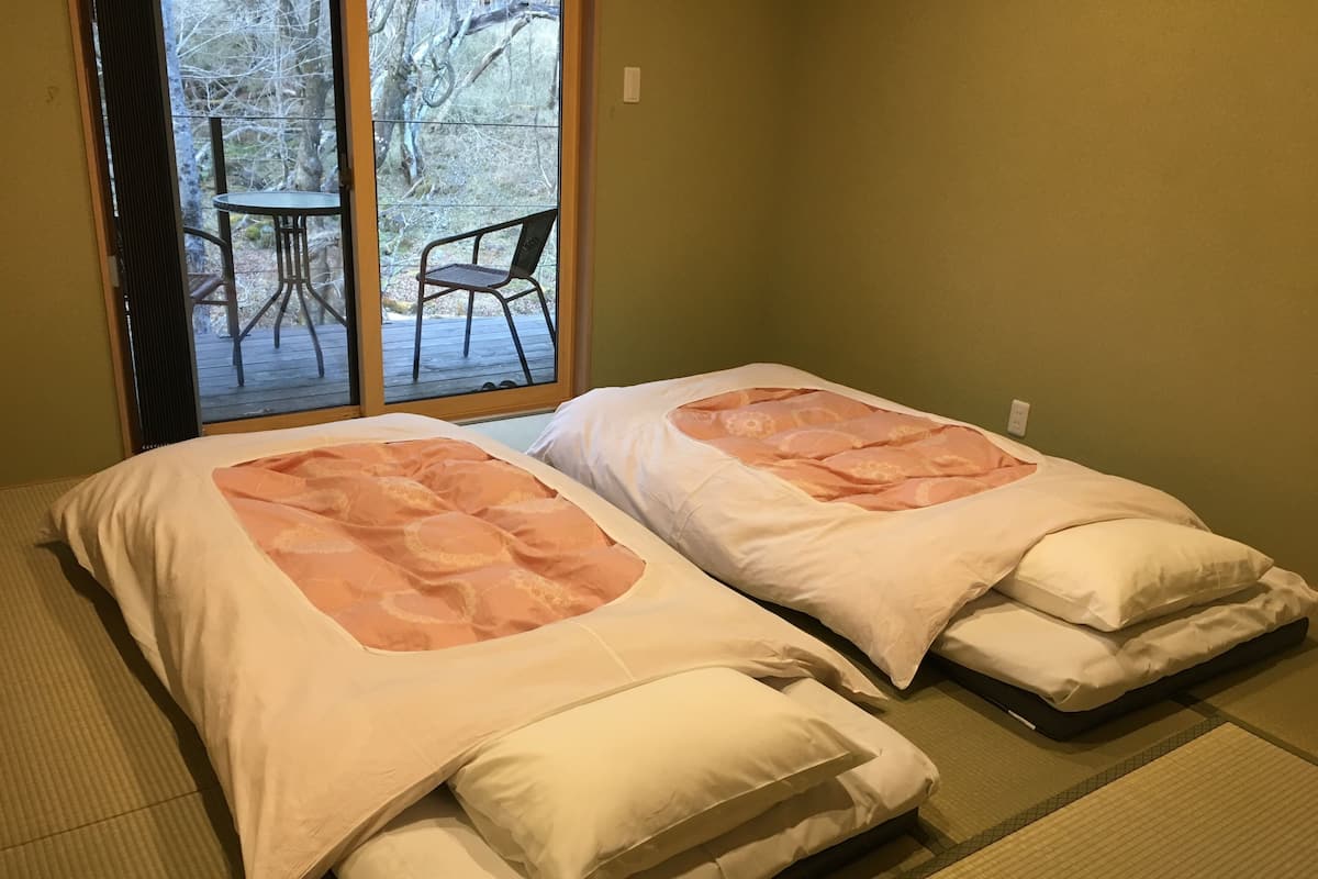Dos camas individuales con ropa de cama blanca y melocotón, una puerta corredera de vidrio que da a un espacio exterior con una silla.