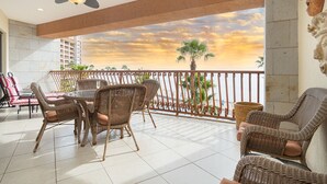 Un balcón con mobiliario de mimbre, un ventilador de techo y vistas a palmeras y un atardecer.
