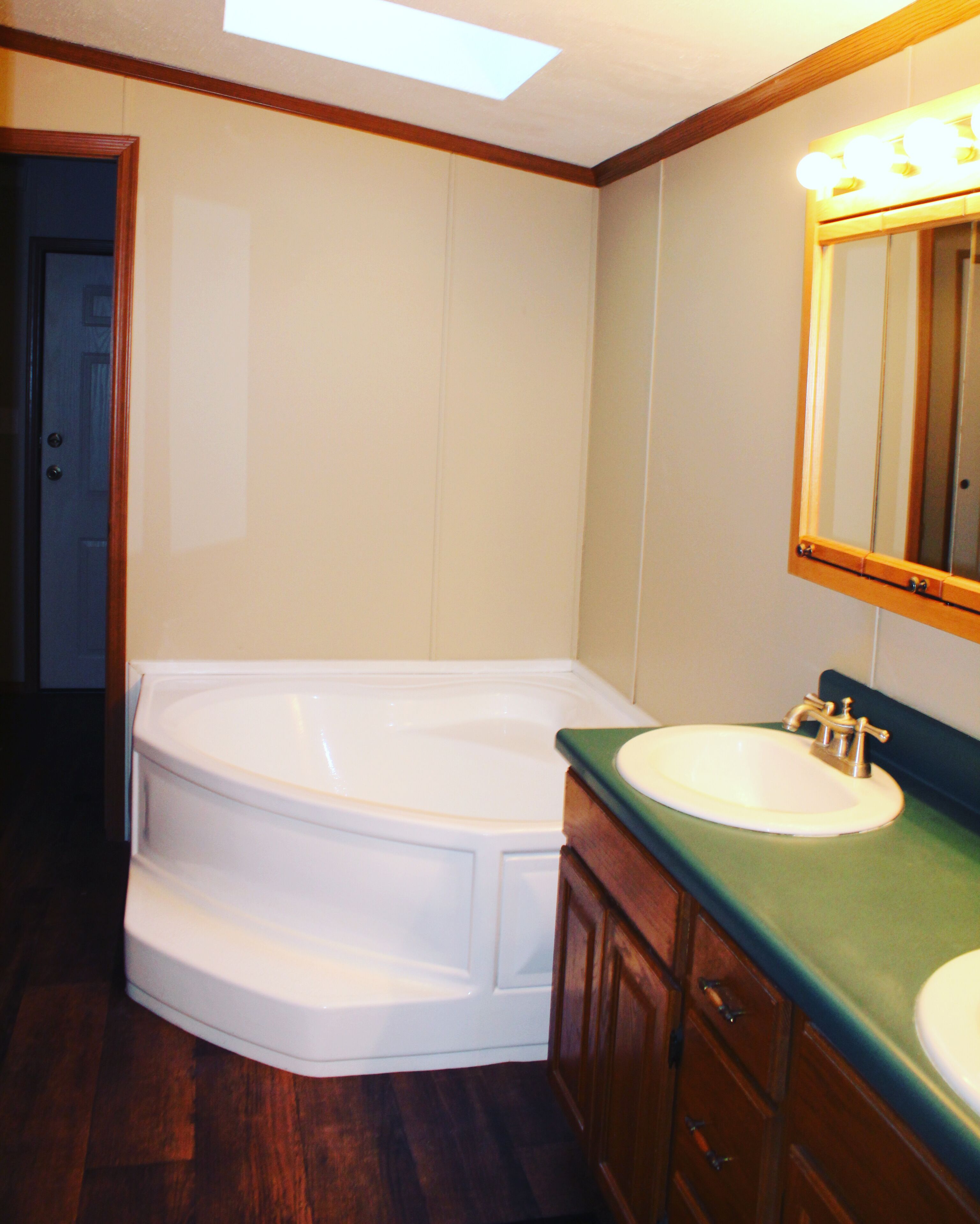 A bathroom with a white bathtub, a sink with a green countertop, wooden cabinets, and a mirror with lights above it.