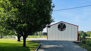 A metal building with a circular logo, a gravel path leading to it, and a large tree to the left. - Secluded Farm near Bennett Springs State Park, 66 Robin Ln Tunas, MO 65764 (Lebanon)