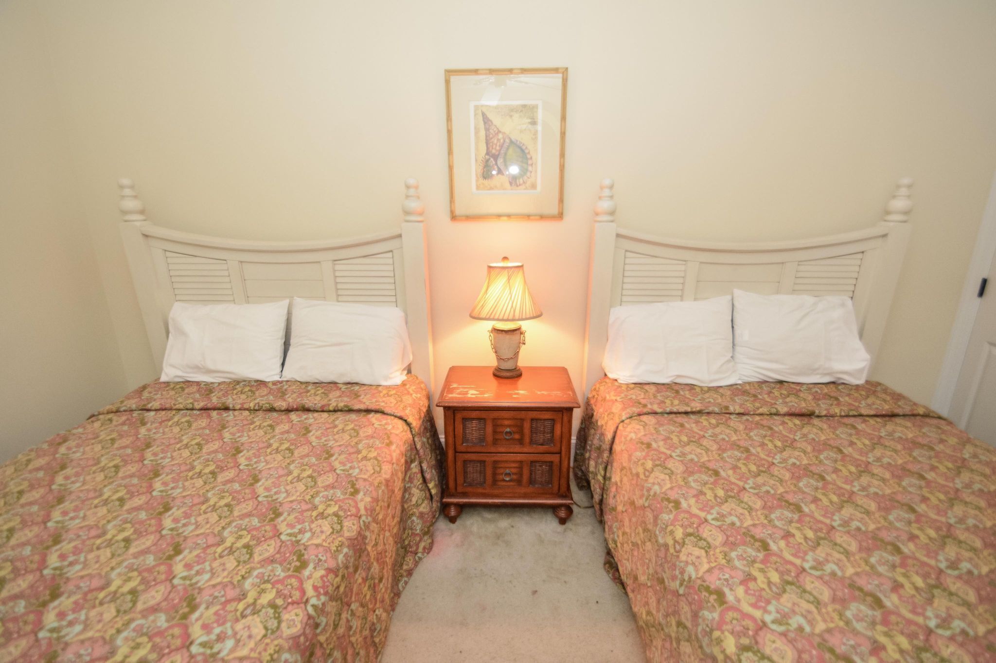Quarto com duas camas, cada uma com colcha estampada, uma mesa de cabeceira de madeira e um abajur, além de um quadro na parede.