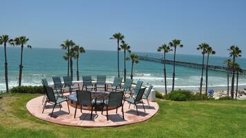 A circular outdoor seating area with chairs and a fire pit, overlooking a beach with palm trees and a pier.