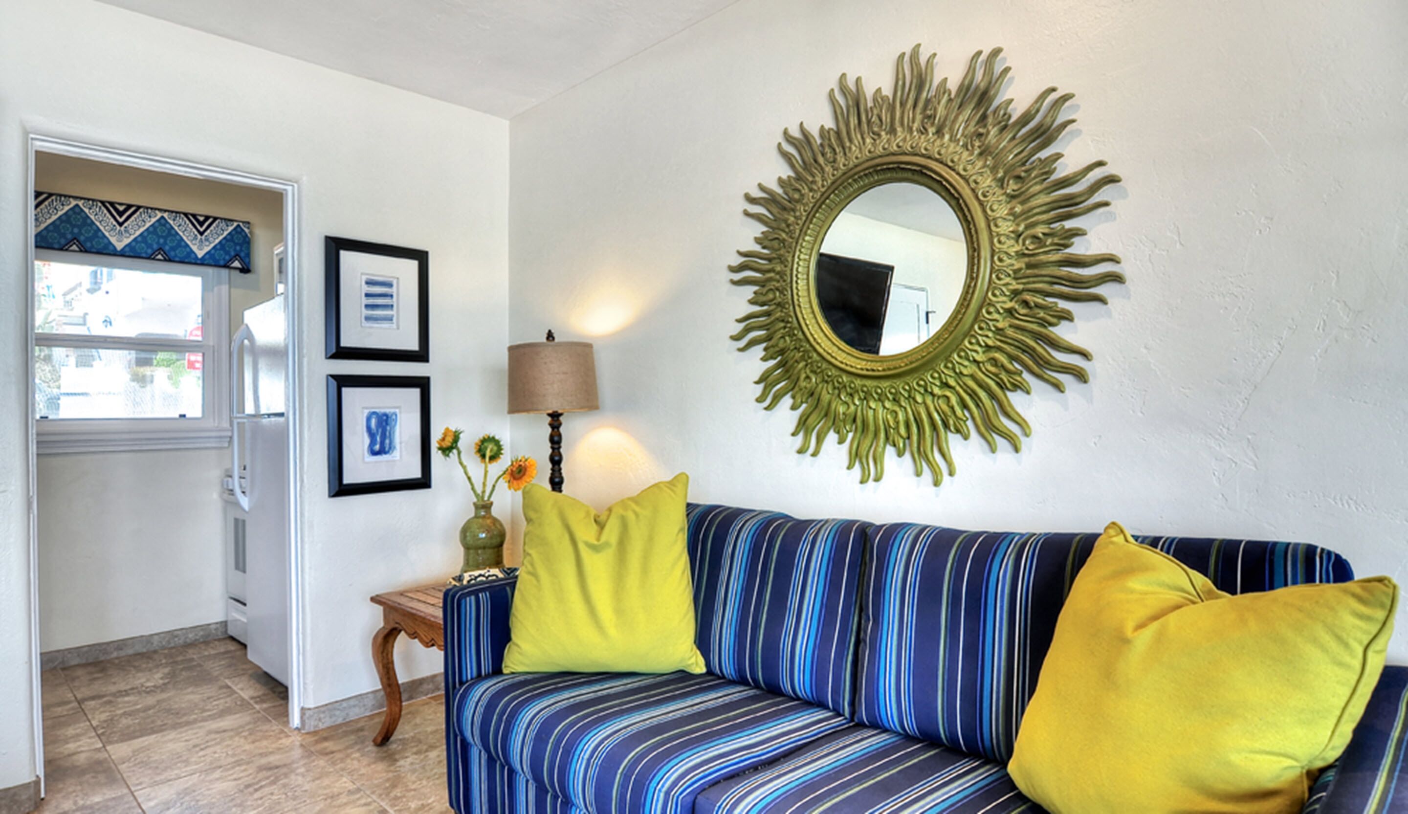 Un salón con un sofá a rayas, dos cojines amarillos, un espejo con diseño de sol y obras de arte enmarcadas en la pared.