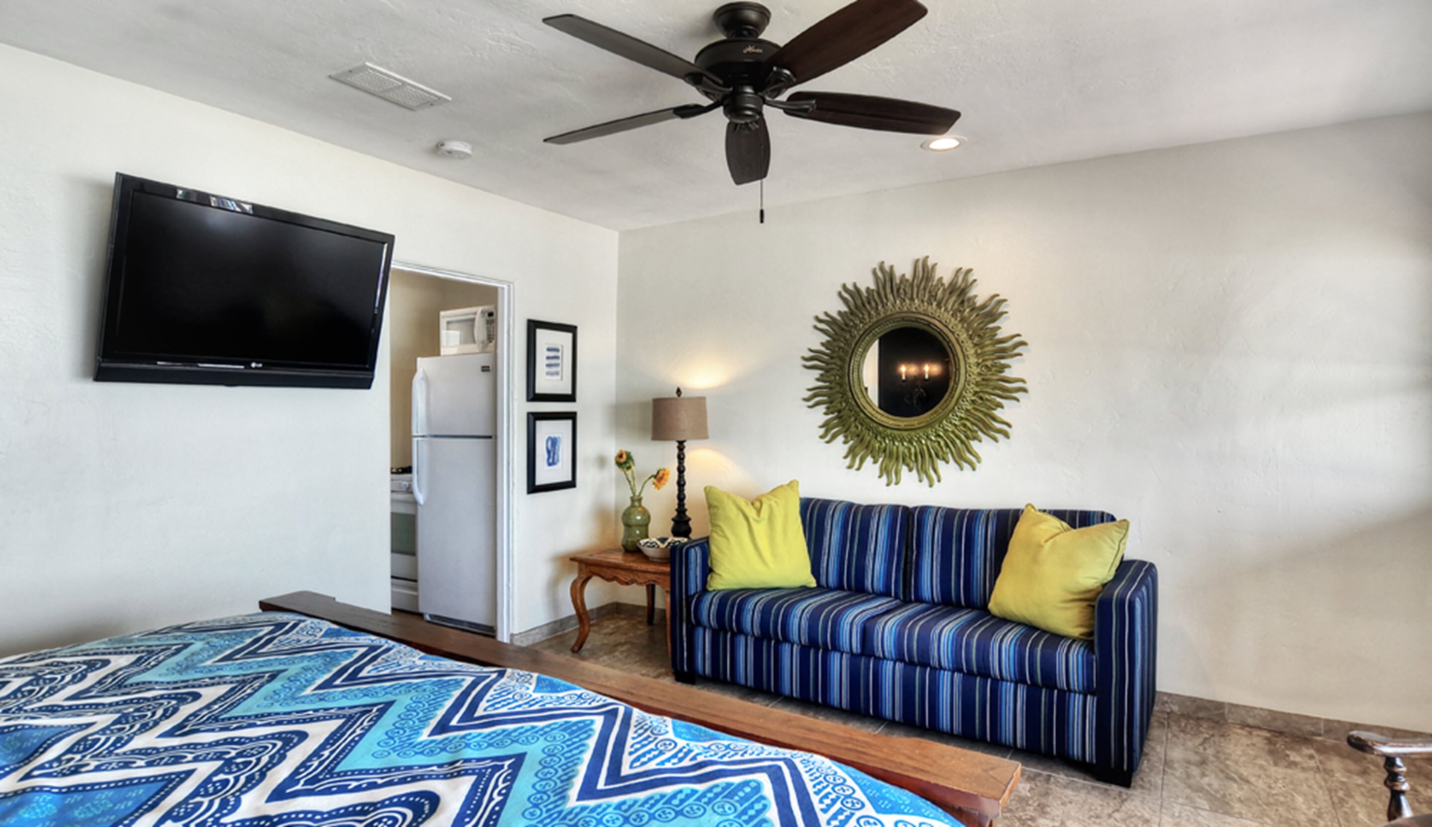 A hotel room with a bed, a sofa, a ceiling fan, a flat-screen TV, a microwave, and a decorative mirror.