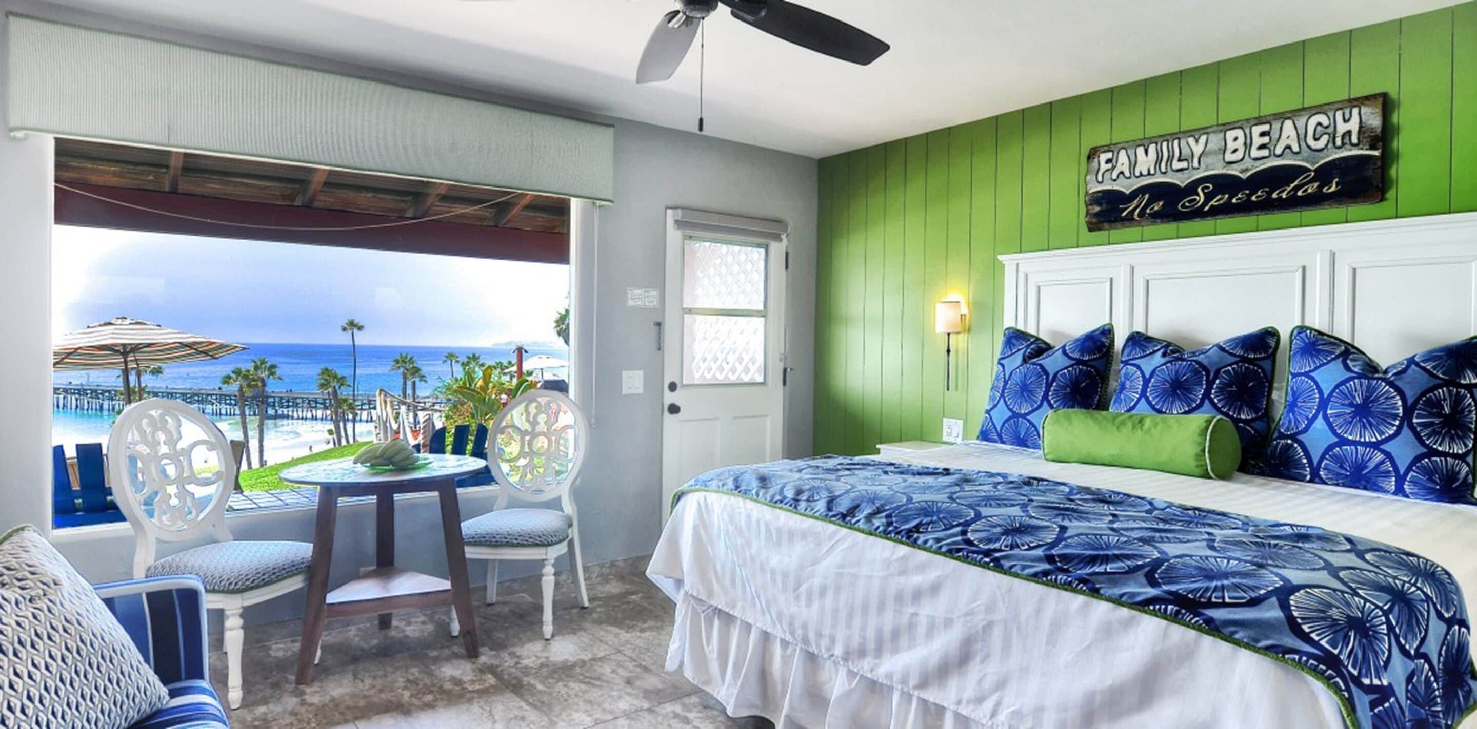 A bedroom with a view of the ocean, a bed with blue floral bedding, and a green wall with a 'Family Beach' sign.