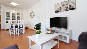 A living room with a white TV stand, a flat-screen TV, a dining area with white chairs and a cabinet, and a potted plant. - 1155 Apartment SanJuan 18 (Nerja)