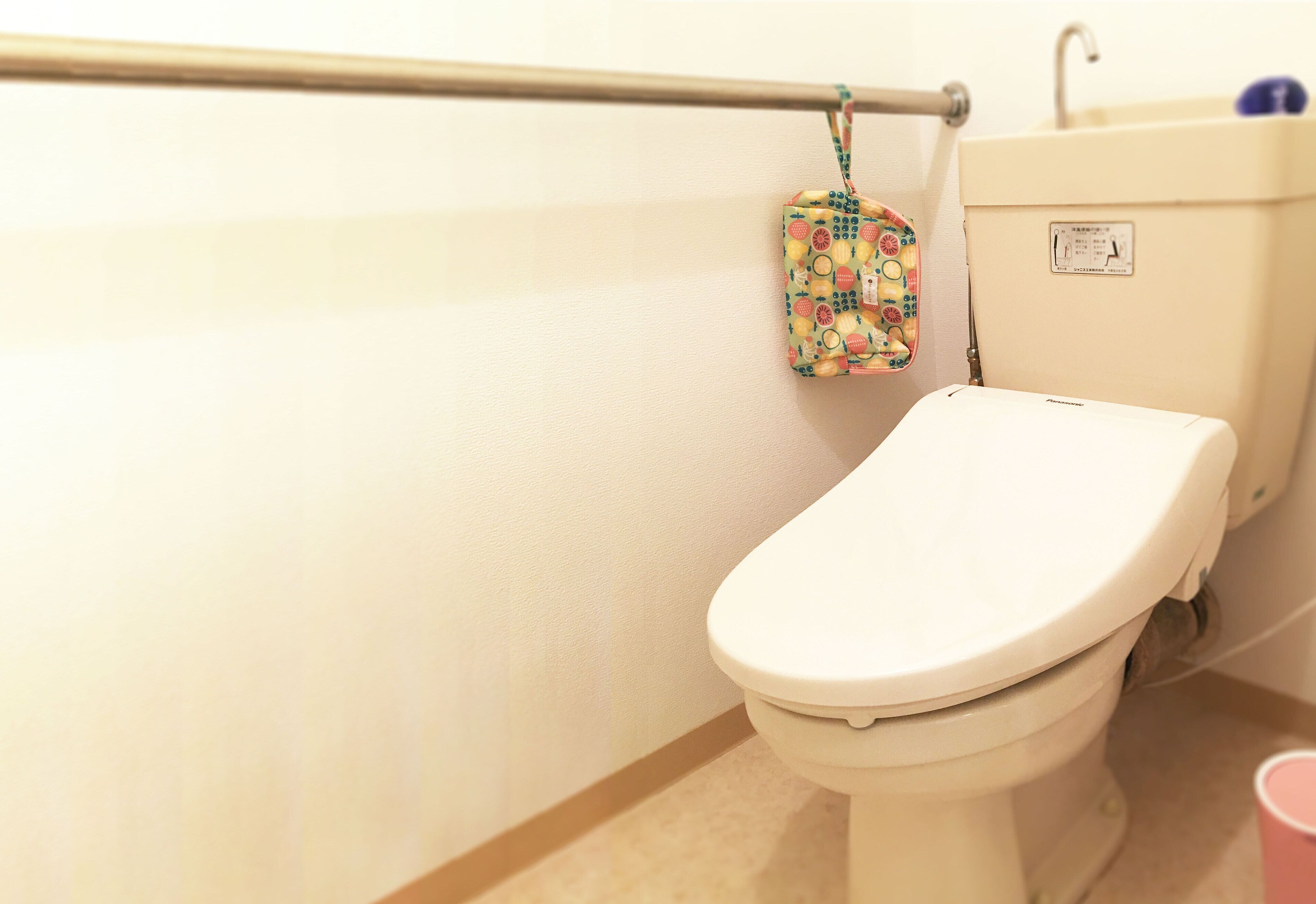 A toilet with a white lid, a wall-mounted handrail, and a small hanging bag with a fruit pattern.
