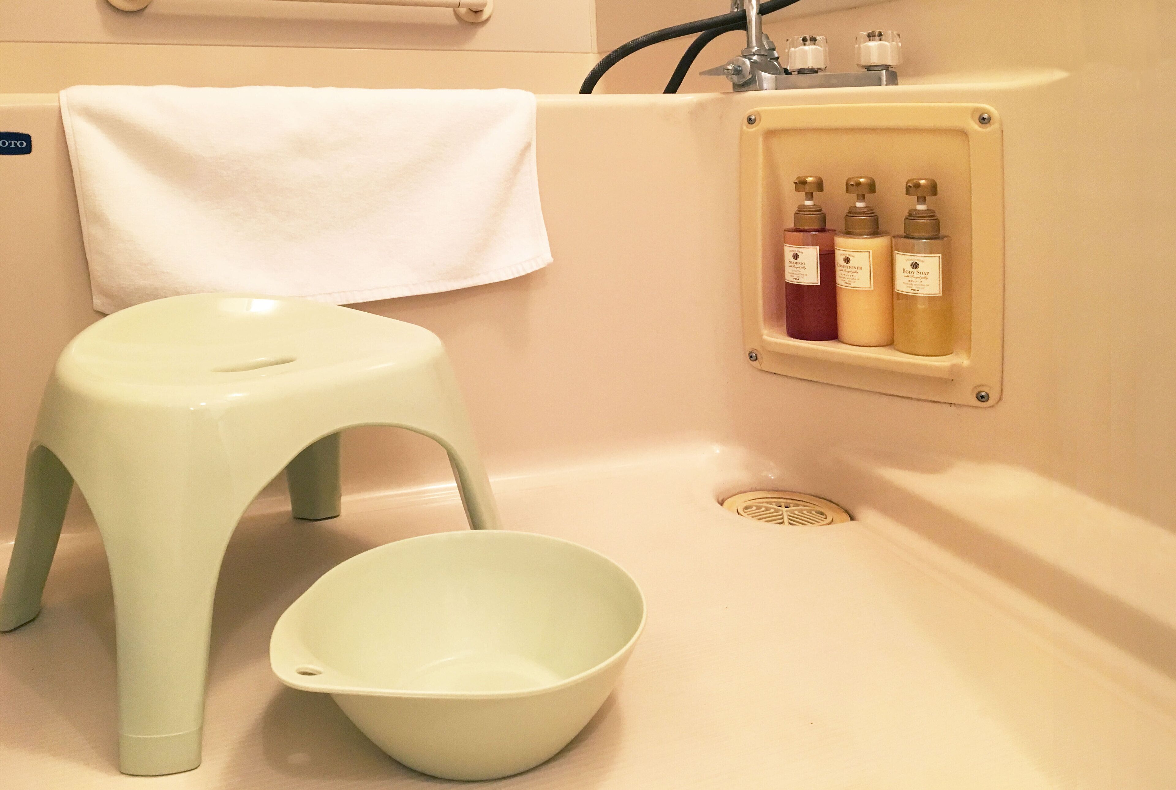 A bathroom with a white bathtub, a white towel hanging on the wall, a green stool, and a shelf with three bottles of toiletries.