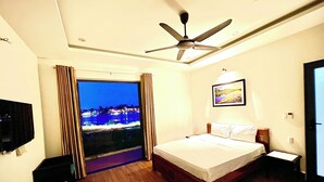 A hotel room with a bed, a ceiling fan, a TV, and a window with a view of a cityscape at night. - Thuong Da Riverside Homestay (Da Nang)