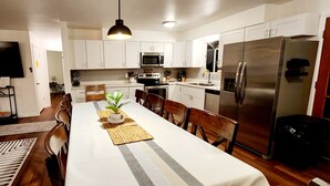 Cozinha moderna com armários brancos, eletrodomésticos de aço inoxidável e uma área de jantar com uma mesa comprida e cadeiras.
