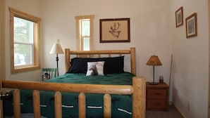 A bedroom with a wooden bed frame, a green bedspread with animal prints, a deer pillow, two framed pictures on the wall, a nightstand with a lamp, and a window with a wooden frame.