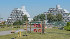 Un parque infantil con una estructura de escalada y columpios, rodeado de un parque con césped y árboles, y edificios modernos al fondo.