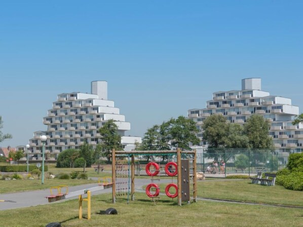 A playground with a climbing structure and swings, surrounded by a park with trees and a road.