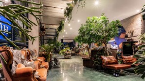A spacious lobby with leather sofas, a large potted plant, and a mural on the wall. - The Wood (Surin)