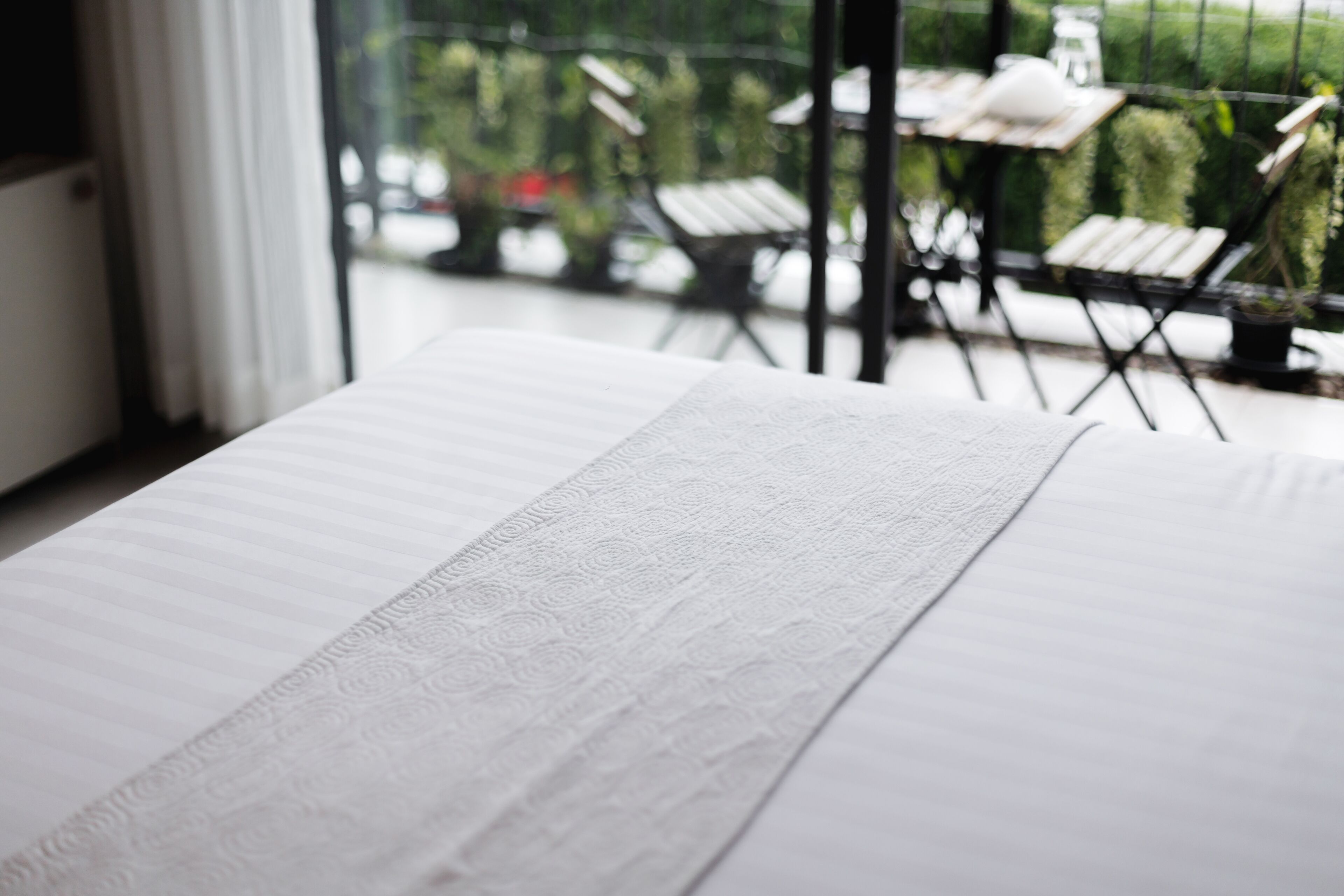 A bed with a textured white sheet, a window with a view of outdoor seating and greenery.