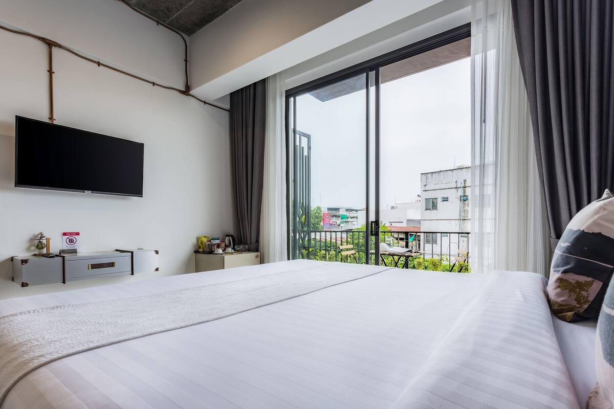 Una habitación de hotel moderna con una cama grande, un televisor de pantalla plana montado en la pared, un escritorio pequeño con una silla y un balcón con vista a la ciudad.