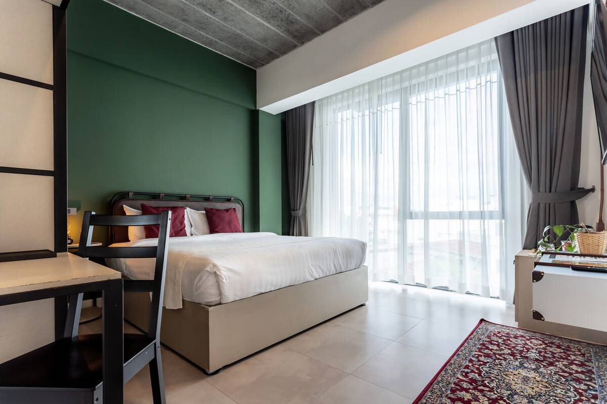 Una habitación de hotel moderna con cama, escritorio y silla. Cuenta con un ventanal con cortinas, una alfombra roja y una planta sobre una mesita de noche.