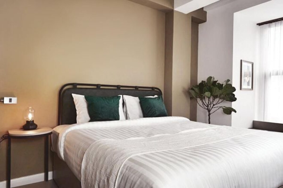A neatly made bed with a headboard, a bedside table with a lamp, and a potted plant in a modern room with neutral walls.