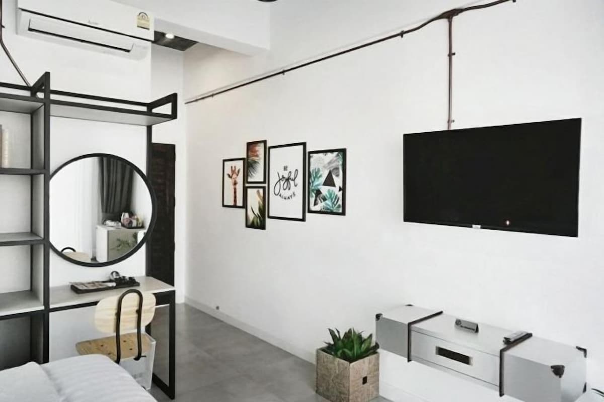 A modern bedroom with a bed, a round mirror, a desk with a chair, a TV mounted on the wall, and framed artwork.
