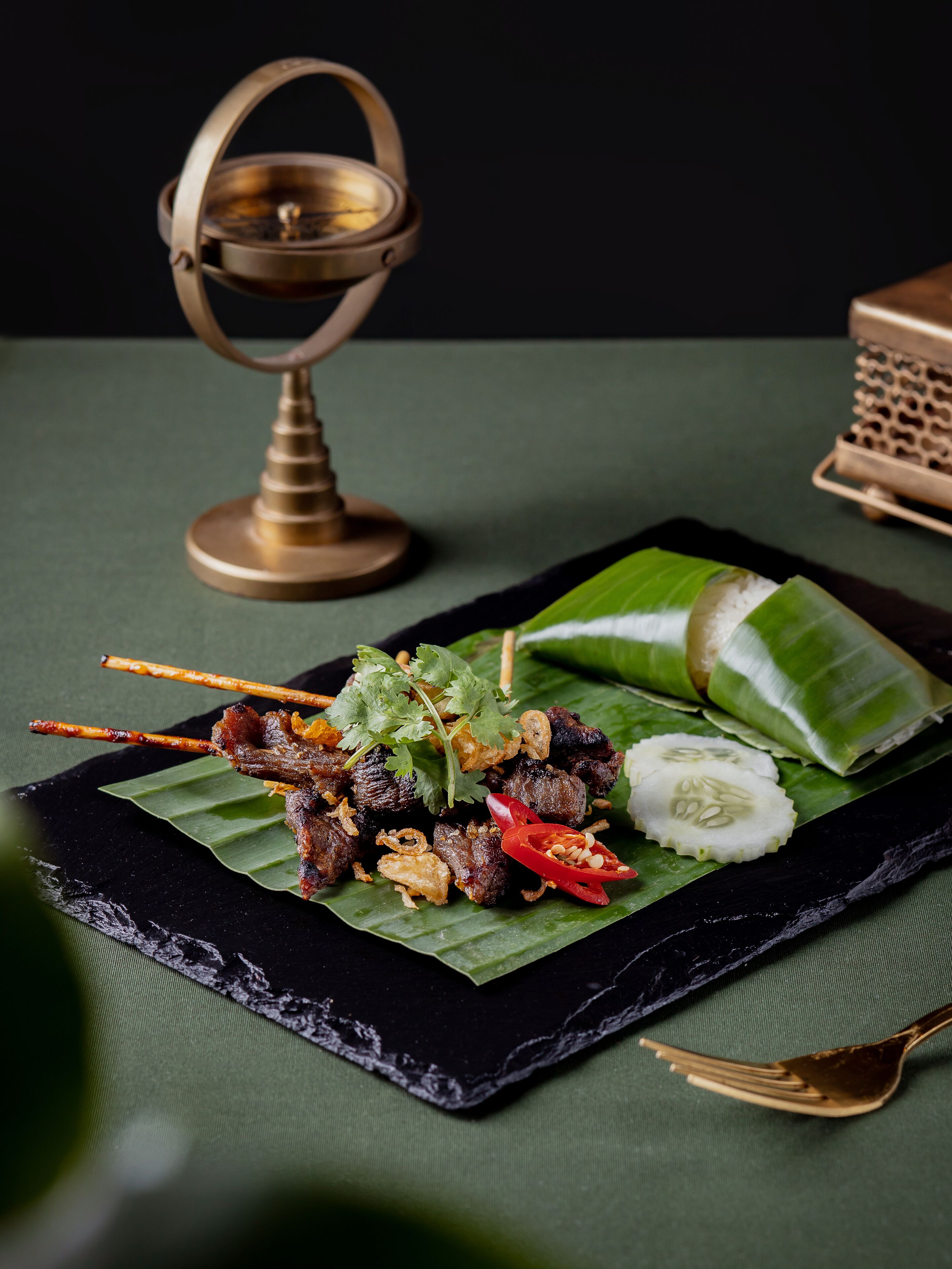 A black slate serving tray with skewers of grilled meat, a slice of cucumber, and a piece of white rice wrapped in banana leaves, set on a green cloth with a gold decorative item in the background.