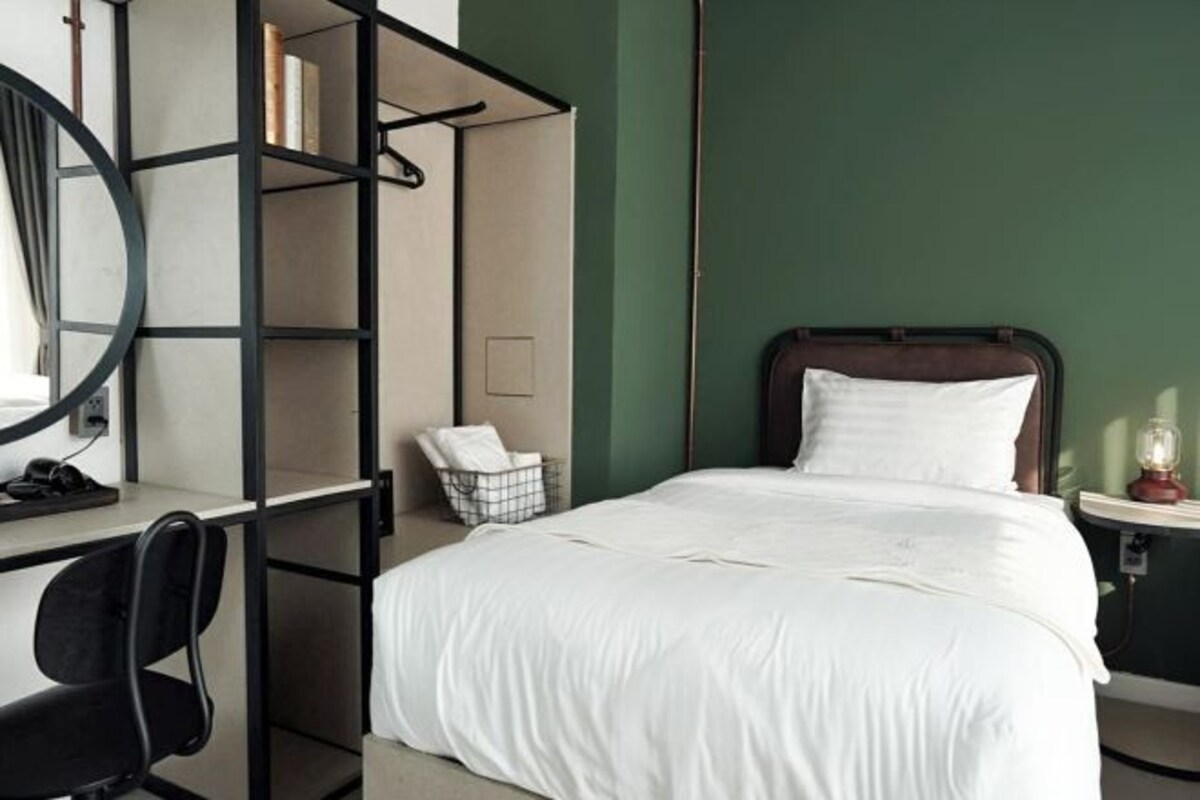 A neatly arranged hotel room with a bed, a desk with a chair, a bookshelf, and a bedside table with a lamp.