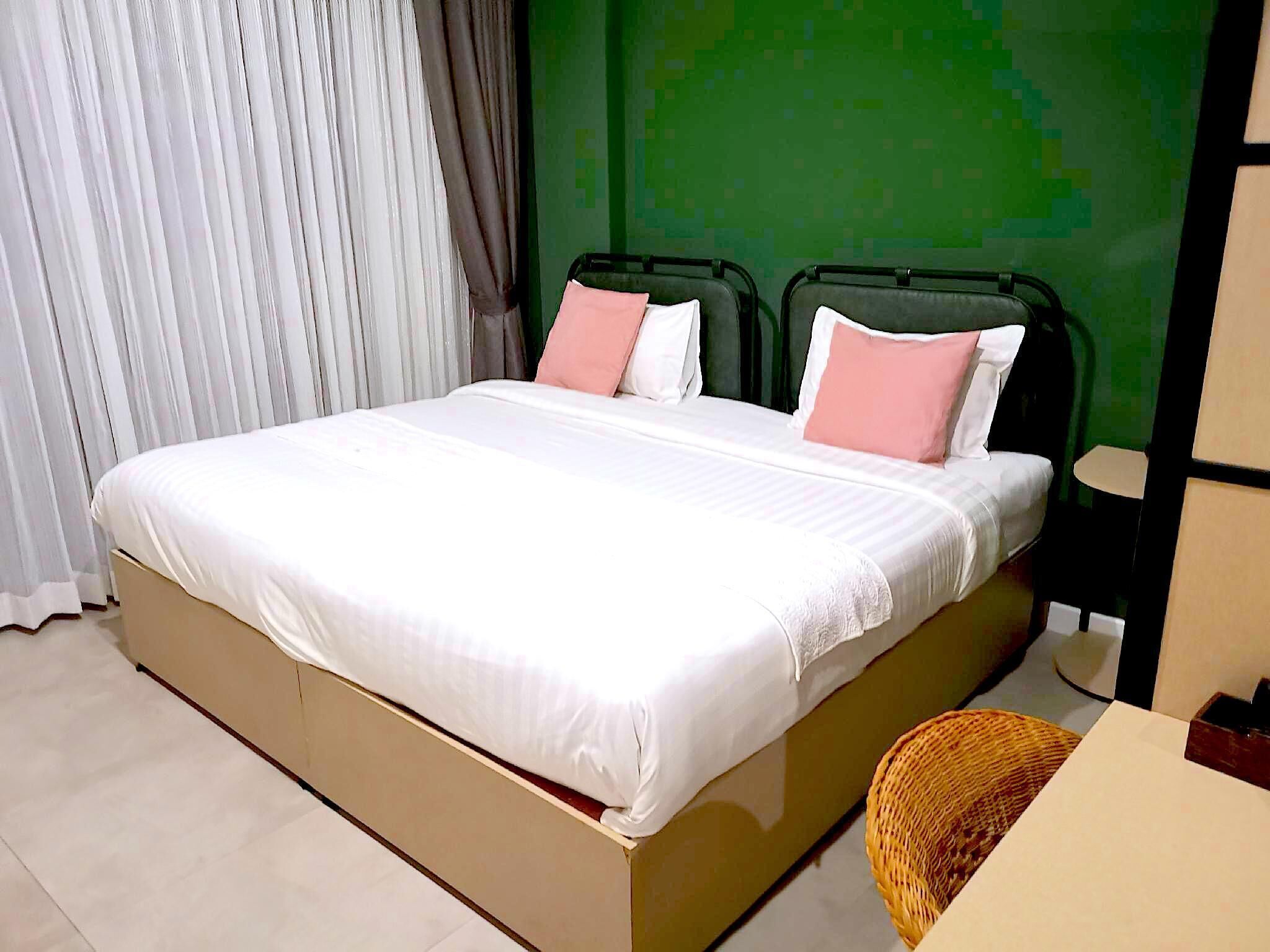 Una cama con ropa blanca y almohadas rosadas, una pared verde y una ventana con cortinas blancas.