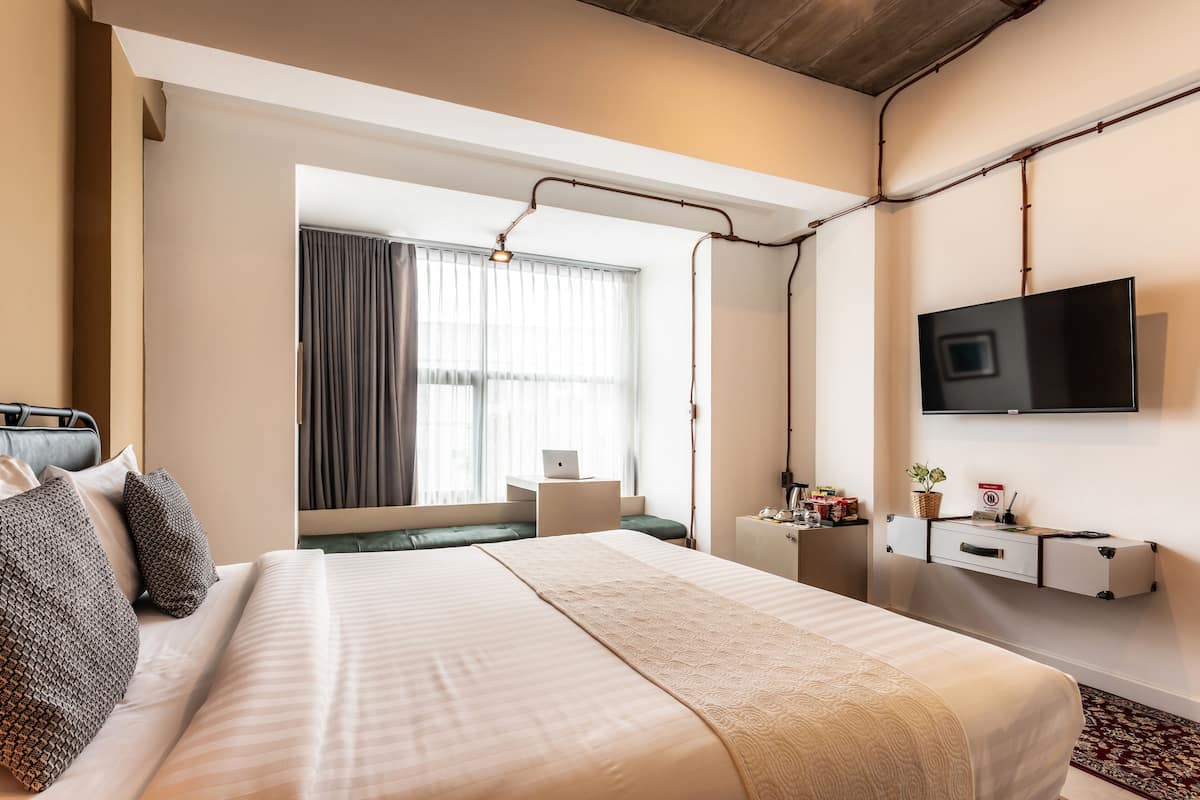 Habitación de hotel moderna con una cama grande, un escritorio con una computadora portátil, un televisor y una pequeña planta en un estante.