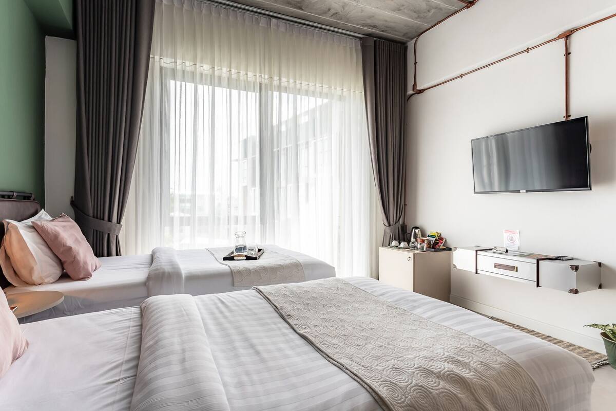 Habitación de hotel con dos camas, televisión, mesita de noche con artículos de aseo y un ventanal con cortinas translúcidas.