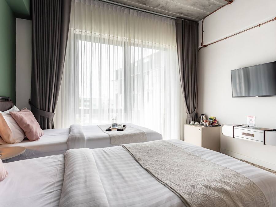Habitación de hotel con dos camas, televisión, mesita de noche con artículos de aseo y un ventanal con cortinas translúcidas.