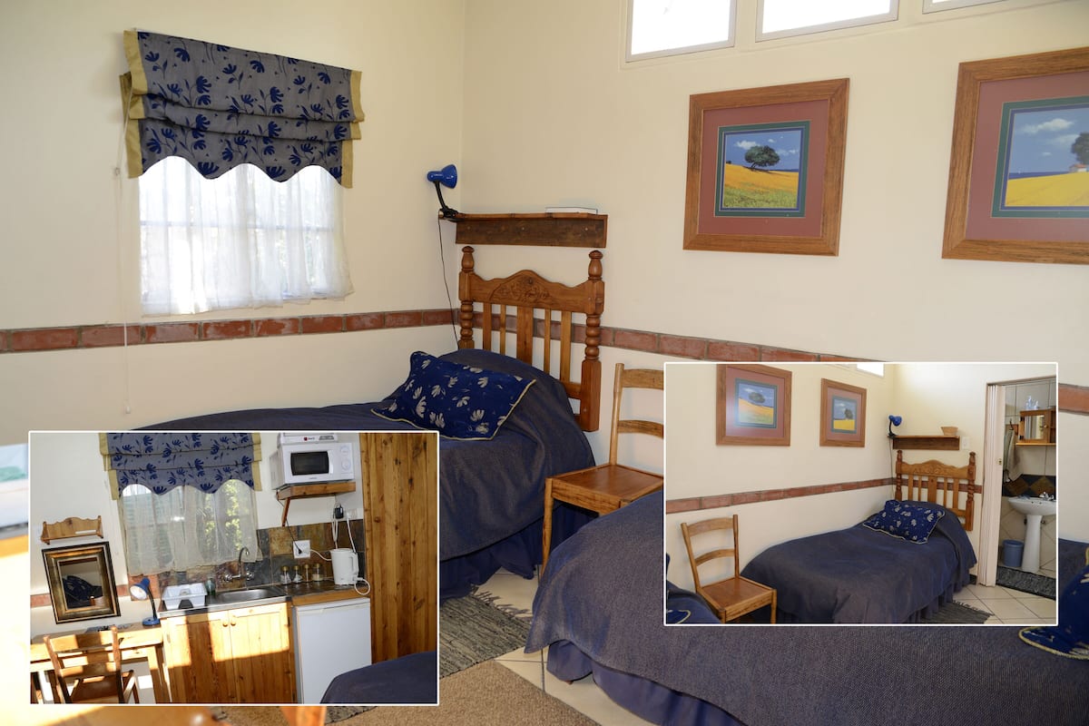 A bedroom with a single bed, a wooden headboard, a nightstand, a desk with a chair, a window with curtains, and a wall with framed pictures.