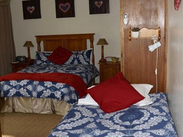 A bedroom with two beds, each with a patterned bedspread and red pillows. There are bedside tables with lamps and a wooden door with a small shelf above it. - Marnicus Guesthouse (Matjhabeng)