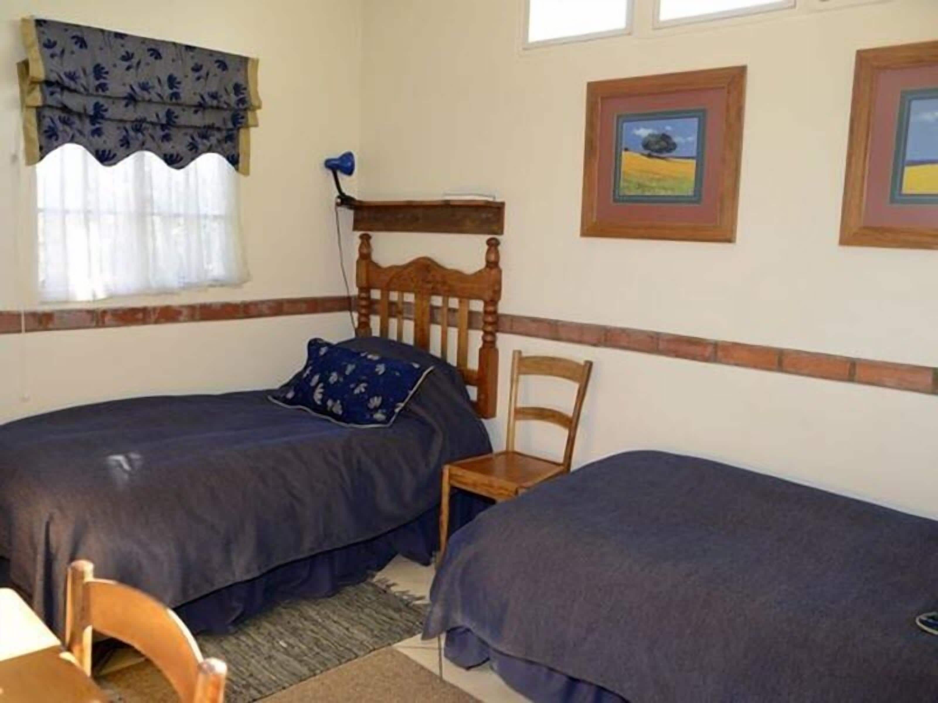Une chambre avec deux lits simples, une tête de lit en bois, une chaise et deux tableaux encadrés accrochés au mur.