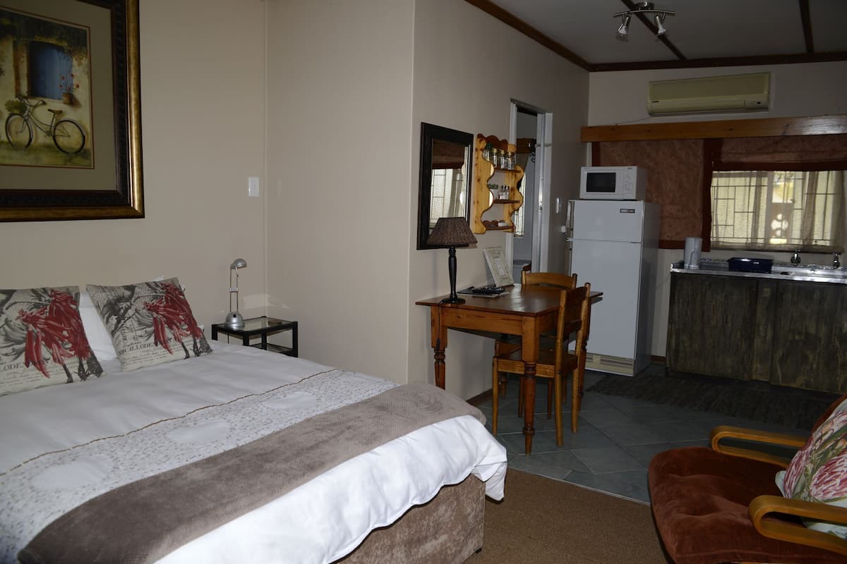 A bedroom with a bed, a desk, and a chair. There is a kitchenette with a refrigerator and a microwave.