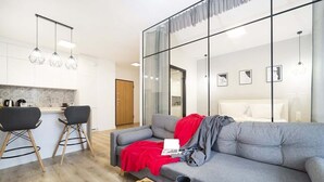 Un salon moderne avec un canapé gris, une table basse sur laquelle on trouve des croissants et une tasse, et un coin cuisine avec des tabourets de bar et des luminaires suspendus.