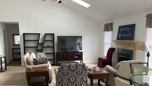 A living room with a sofa, armchair, ottoman, and a glass table. There is a fireplace with a sign above it, and a television on a stand. - A Den for all four seasons, visit the Bear's Den and create lasting memories. (McGaheysville)