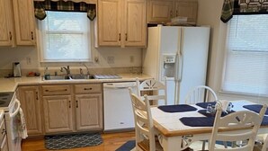 A compact kitchen with wooden cabinets, a white refrigerator, a dining table with chairs, and a blue rug. - A Den for all four seasons, visit the Bear's Den and create lasting memories. (McGaheysville)