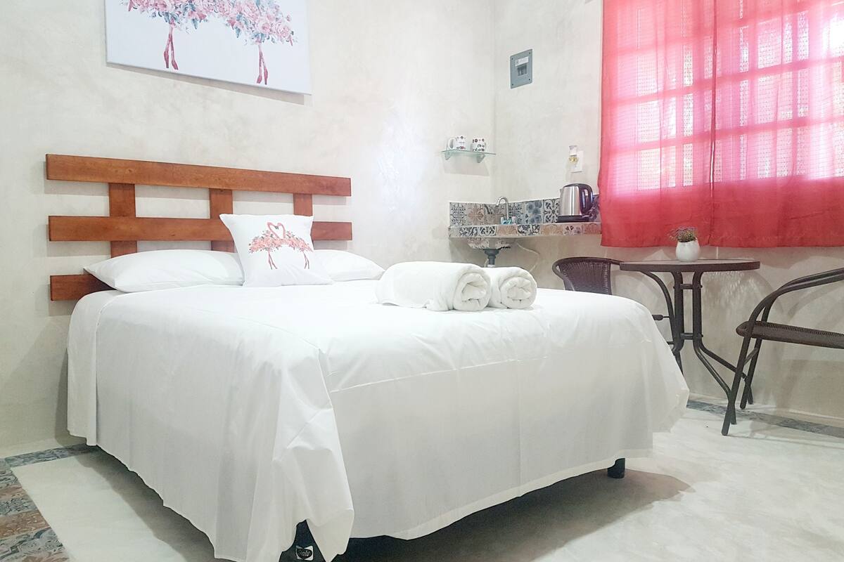 Une chambre d’hôtel avec une tête de lit en bois, un lit recouvert de draps blancs, une œuvre d’art représentant un flamant rose accrochée au mur, une petite table avec une bouilloire et un rideau rouge.
