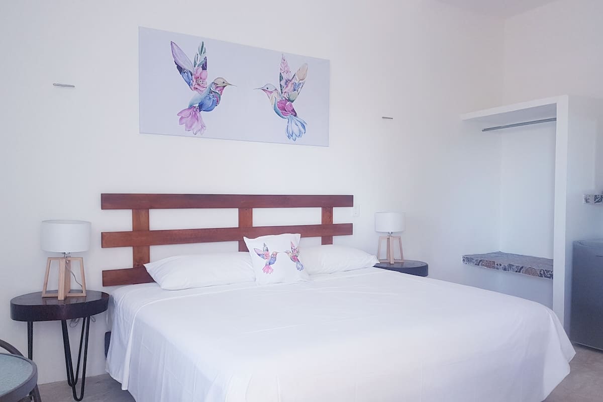 Une chambre à coucher avec un grand lit, deux tables de chevet, une tête de lit en bois et un tableau représentant des oiseaux, accroché au mur.