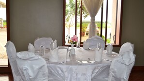 A dining table set for six with white tablecloth and chairs, a vase with pink flowers, and a view of the outdoors through a window. - Panone Hotel Chekereni (Arusha)
