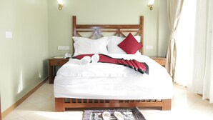 A neatly made bed with white linens, a red pillow, and a blanket. There are two bedside tables with lamps, a phone, and a folded towel. The room has light green walls and a wooden headboard. - Panone Hotel Chekereni (Arusha)