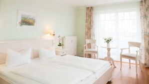 A hotel room with a large bed, two bedside tables, a chair, a small table, and a window with curtains.