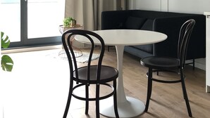 Un salon moderne avec une table ronde blanche, deux chaises noires, un canapé noir, un lampadaire et une grande fenêtre avec des rideaux.