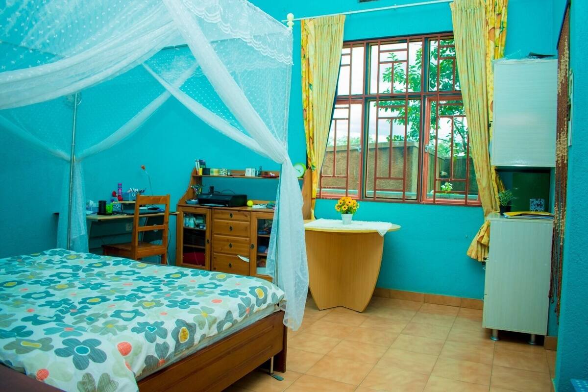 Ein Schlafzimmer mit einem Bett, einem Schreibtisch, einem Stuhl, einem kleinen Tisch mit Blumen und einem Fenster mit Vorhängen.