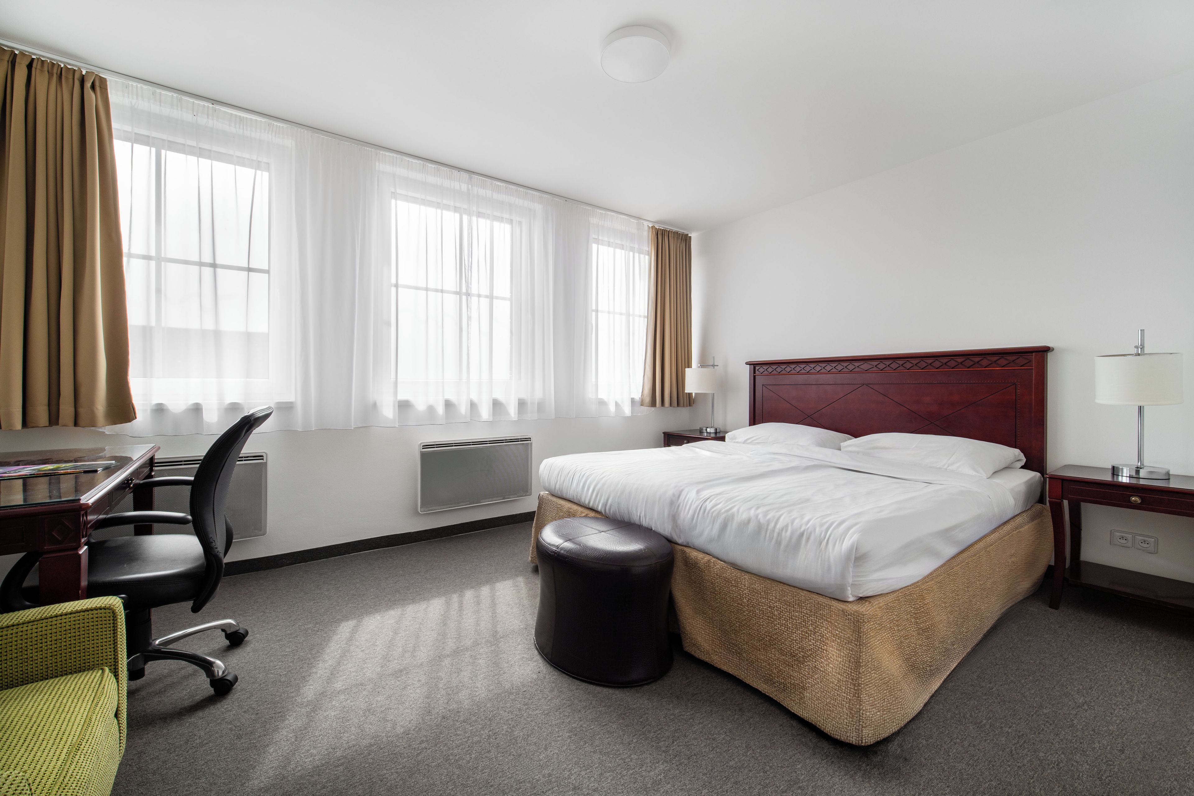 A hotel room with a bed, a desk with a chair, a lamp, and a window with curtains.