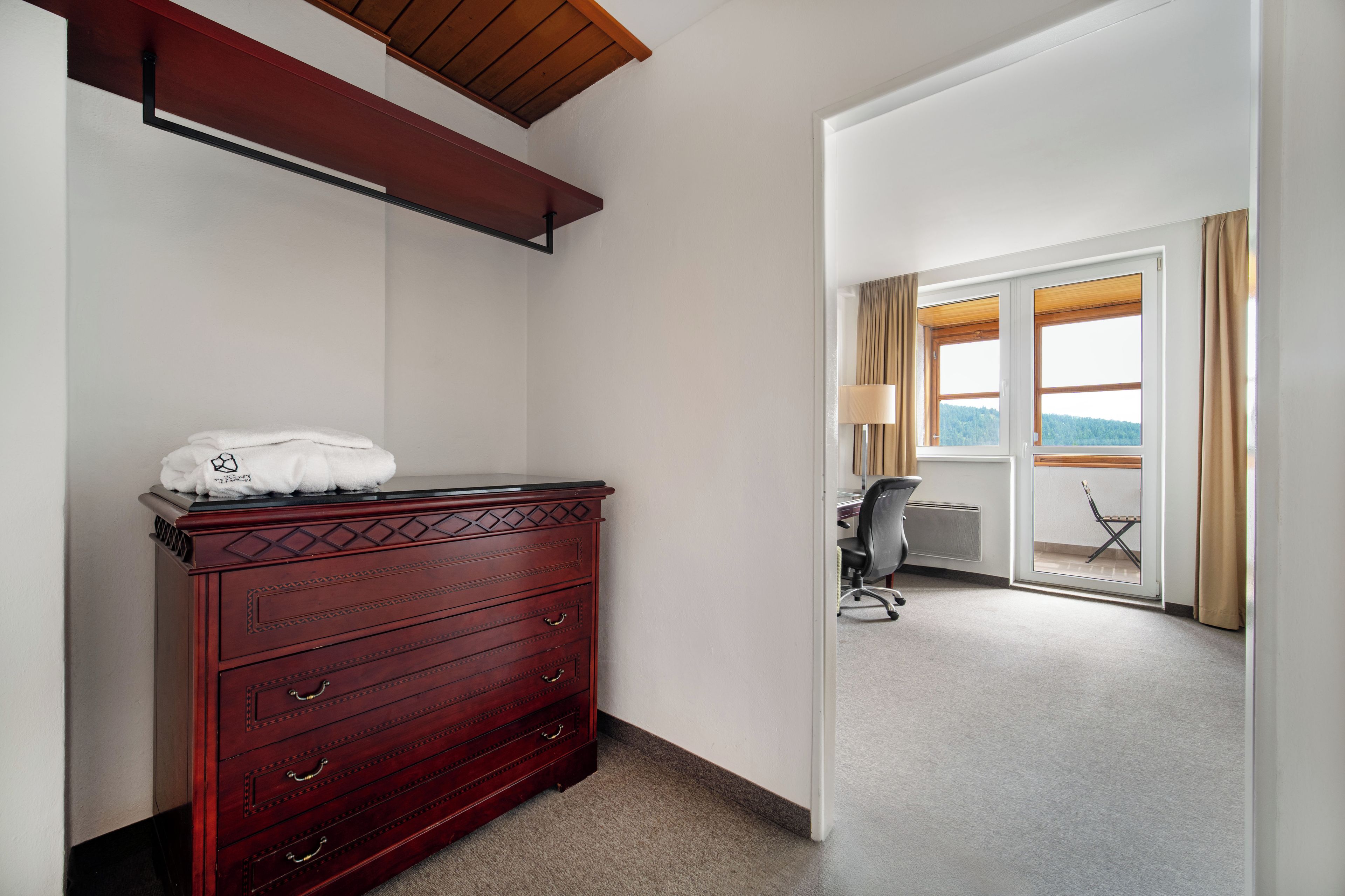 A hotel room with a wooden dresser, a chair, a desk, and a view of the outdoors through a large window.