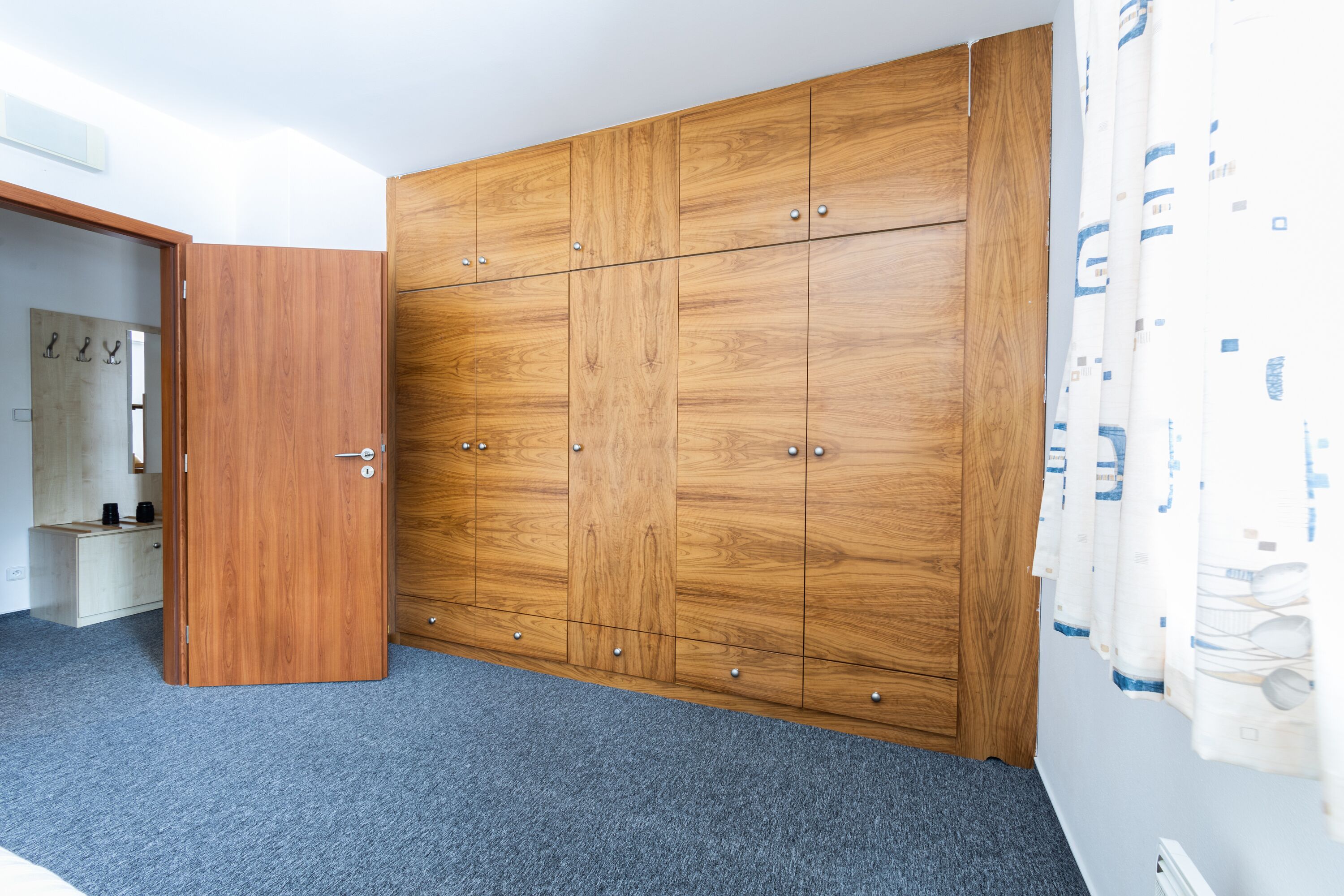 Een kamer met een grote houten garderobe, een grijze vloerbedekking en een gedeeltelijk zichtbare badkamer met spiegel en handdoekrek.