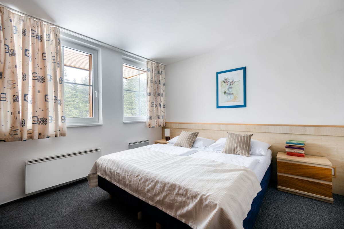 A hotel room with a large bed, a bedside table with books, a framed picture on the wall, and a window with curtains.