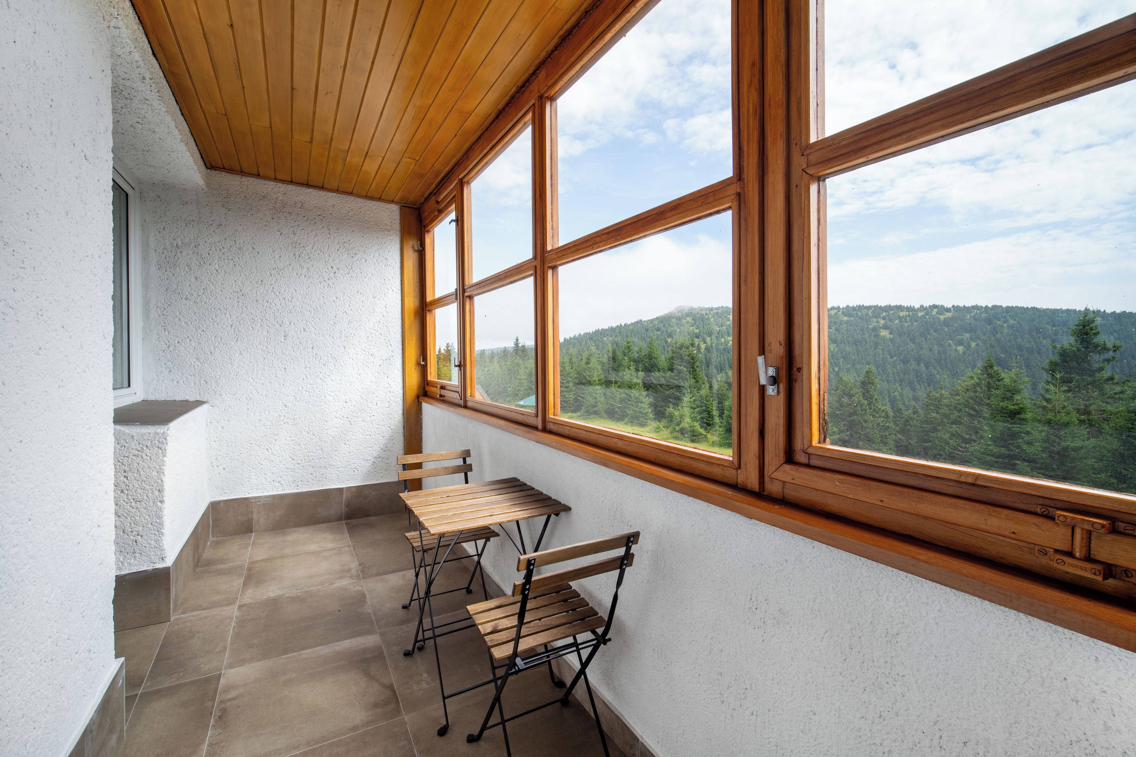 A balcony with a wooden table and chairs, a view of a forested hillside, and a white textured wall.